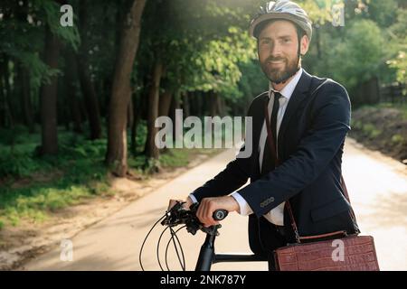 Confident businessman in helmet walking to work with bicycle in morning. Portrait of bearded entrepreneur in stylish suit standing with bike, while lo Stock Photo
