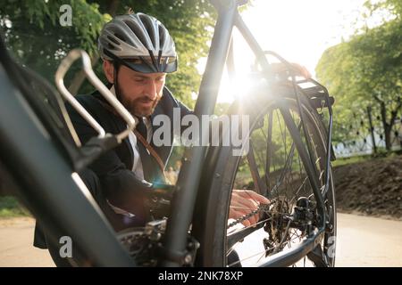 Young bearded businessman checking bicycle before riding to work in morning. Front view of handsome male employee in helmet inspecting bike wheel, whi Stock Photo