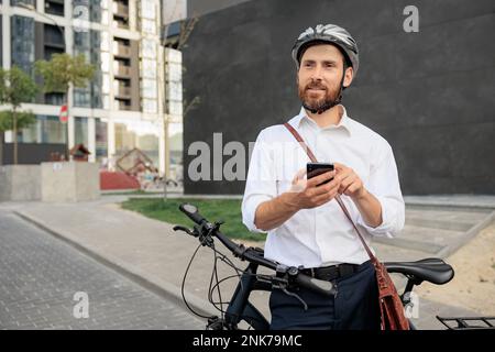 Happy bearded man using online map on smartphone, while checking way to job in city. Front view of smiling male employee in shirt relying on parked bi Stock Photo