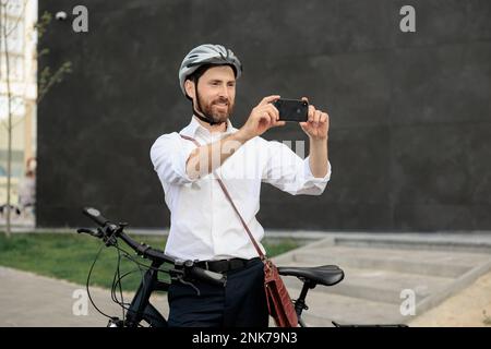 Attractive bearded man taking photo, while standing with bike on street. Front view of smiling male businessman in helmet using smartphone to take pic Stock Photo