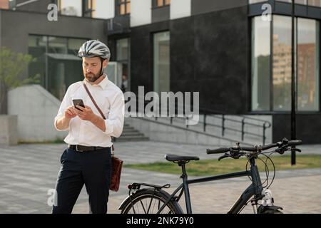 Serious young manager with bike, surfing internet on smartphone, while standing on street. Front view of attractive bearded man in helmet using cell p Stock Photo