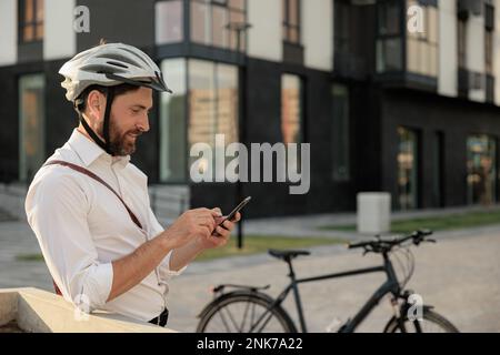 Smiling bearded man with parked bike telephoning, while standing on street. Side view of attractive guy in white shirt using cell phone after working Stock Photo