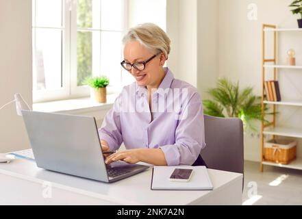 Positive mature elderly woman teacher learn students via Internet in laptop sits in home office Stock Photo