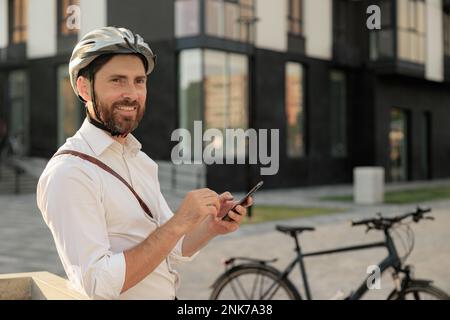 Happy bearded man with parked bike making call, while standing on street. Portrait of attractive guy in helmet using mobile phone, while looking ahead Stock Photo