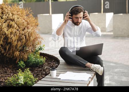 Handsome office worker in white shirt sitting on wooden bench with laptop near documents and coffee cup. Concentrated man wearing headphones and prepa Stock Photo