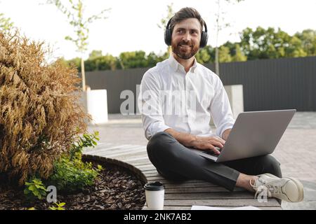 Joyful freelancer sitting on wooden bench with portable laptop on knees. Handsome man in black headphones looking aside with smile on face while worki Stock Photo