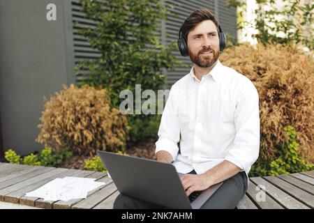Handsome freelancer looking aside while working on modern laptop outdoors. Friendly man in headphones thinking on new project while sitting on wooden Stock Photo