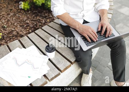 Businessman in white shirt and black pants typing on modern laptop and working hard outdoors. Documents with diagrams and coffee-to-go lying on wooden Stock Photo