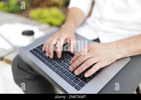 Cropped view of man sitting outdoors while using modern laptop for distance work. Male freelancer in black pants typing emails and touching computer k Stock Photo