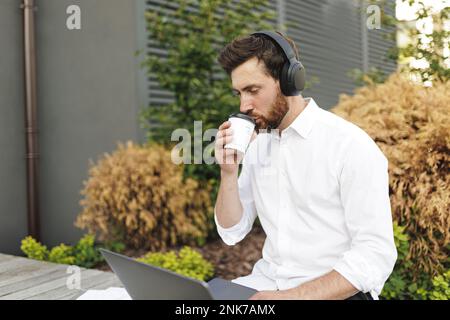 Handsome male freelancer enjoying coffee-to-go and using modern laptop while sitting on wooden bench. Focused man listening music in wireless headphon Stock Photo