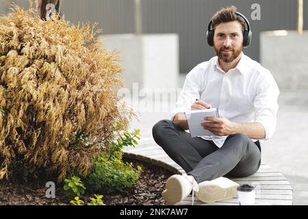 Handsome businessman holding diary in hand and writing down creative ideas. Confident male in headphones looking at camera while sitting on wooden ben Stock Photo