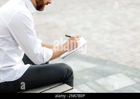 Cropped view of bearded businessman sitting on bench in urban area while making some notes. Stylish male in formal clothing writing down creative idea Stock Photo