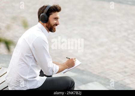 Happy businessman writing new ideas in diary while listening music in headphones. Smiling freelancer with bristle sitting outdoors in quiet place and Stock Photo
