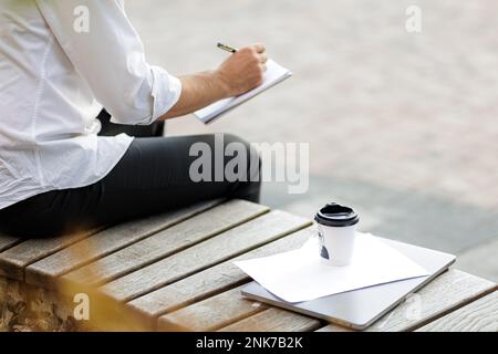 Cropped view of businessman sitting in urban area while writing business plan in notebook. Coffee cup, documents and portable laptop lying near on woo Stock Photo