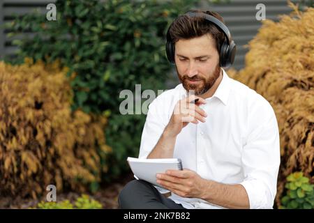 Serious businessman looking on notebook and touching chin with hand in thoughtful gesture. Focused male listening music in wireless headphones while s Stock Photo