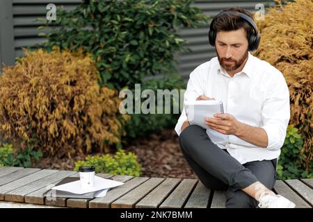 Concentrated bearded man in headphones taking notes, while sitting on bench outdoors. Front view of thoughtful handsome businessman writing, while hav Stock Photo