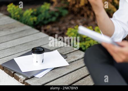 Laptop, notes and paper cup of tasty coffee on wooden bench outdoors. Crop view of unrecognizable entrepreneur working remotely, taking notes during c Stock Photo