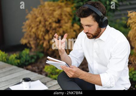Thoughtful man in headphones checking notes, while sitting on bench outdoors. Monochrome portrait of pensive guy with hand on head, thinking, looking Stock Photo
