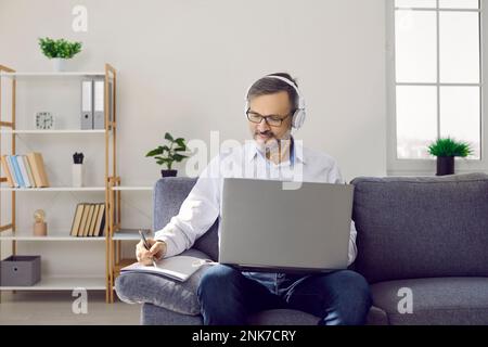 Mature man in headset with laptop working in his home office making notes sitting on couch. Stock Photo
