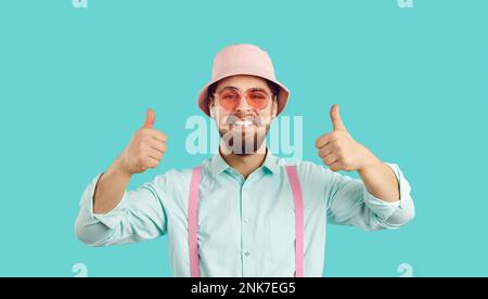 Happy young man wearing pink sunglasses and summer hat smiles and shows thumbs up Stock Photo