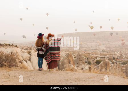 Traveling women wearing authentic boho chic style poncho, sweeter and hats looking on air ballons in sky in Cappadocia valley. Travel and wanderlust c Stock Photo