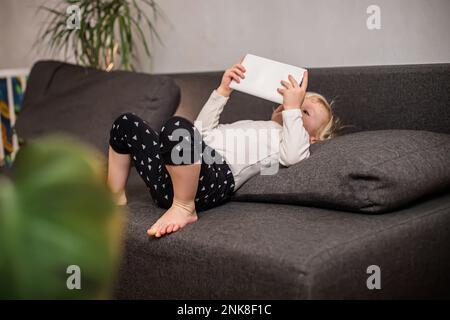 A little girl in a home suit lies on a dark sofa in the room and holds a white tablet in her hands. The child watches cartoons on the gadget. Stock Photo