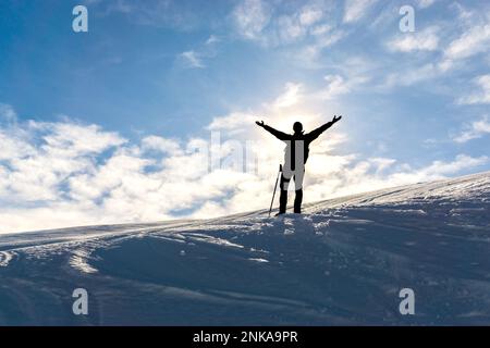 Silhouette of celebrating happy hiker man with raised arms on snowy winter mountain. Sun behind Person. Stock Photo