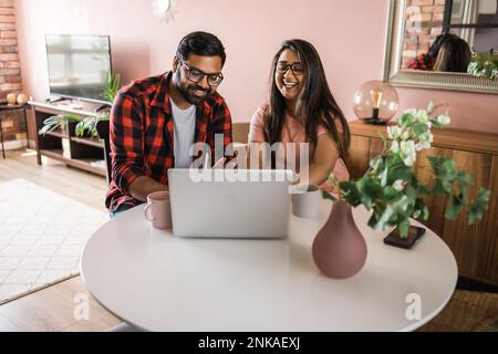 Happy indian family couple cuddle at desk make video call to friends using laptop webcam. Loving young spouses look at computer screen waving hands in Stock Photo