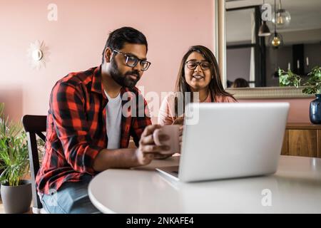 Happy indian family couple cuddle at desk make video call to friends using laptop webcam. Loving young spouses look at computer screen waving hands in Stock Photo
