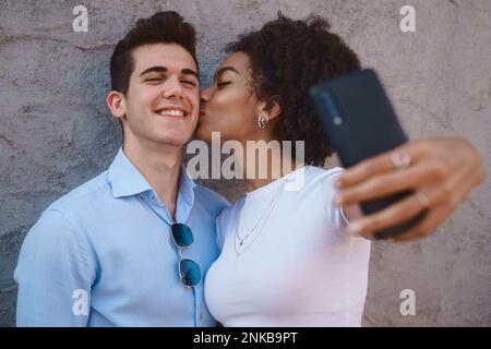 Multiracial couple taking a selfie, a young African American woman with curly hair kissing a Caucasian man on the cheek against a cement wall. Love, i Stock Photo