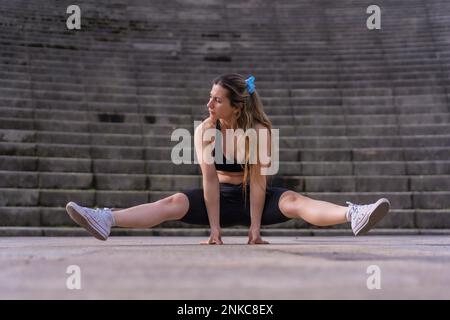 Caucasian young woman doing fitness in a city park, doing the splits and rising up in the air Stock Photo