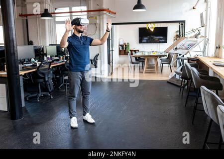 Young businessman wearing virtual reality simulator standing with hands raised in office Stock Photo