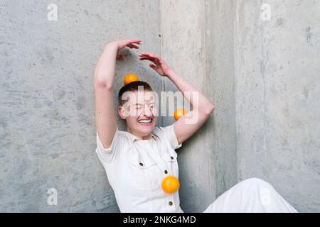 Cheerful non-binary person having fun with oranges in front of wall Stock Photo