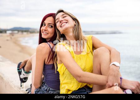 Happy friends sitting back to back at promenade by sea Stock Photo