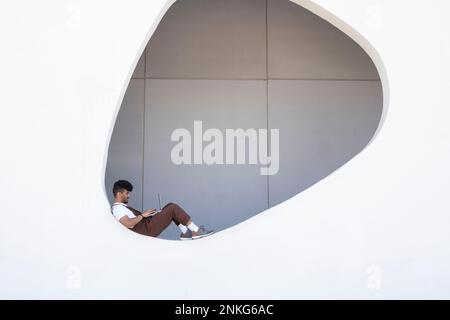 Man using laptop sitting on wall with hole in building Stock Photo