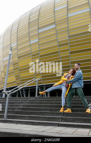Playful woman falling over man on steps in front of stadium building Stock Photo