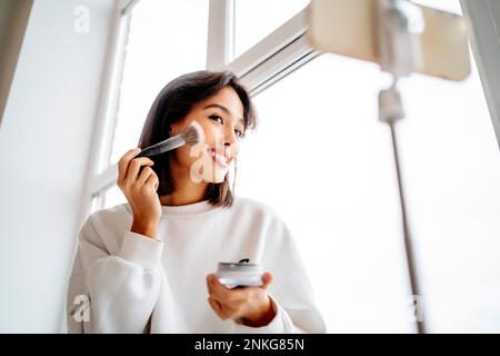 Young influencer applying blusher and filming through mobile phone at home Stock Photo