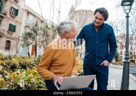 Smiling son with father holding laptop sitting by plants Stock Photo