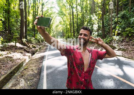 Happy man showing peace sign gesture and taking selfie in front of trees Stock Photo
