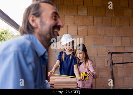 Happy man with daughters measuring bricks in front of wall Stock Photo