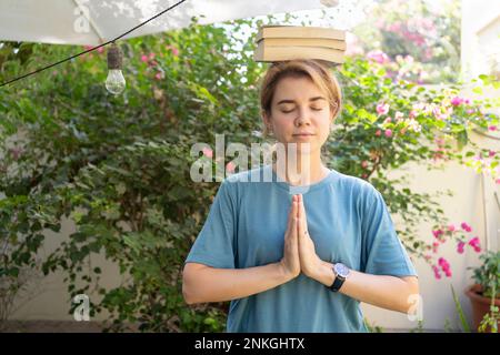 Woman with eyes closed and hands clasped balancing books on head Stock Photo