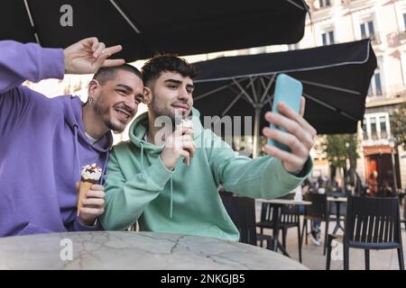 Happy gay couple with ice cream taking selfie through smart phone at cafe Stock Photo