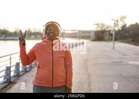 Smiling senior woman wearing headphones showing peace sign at promenade Stock Photo