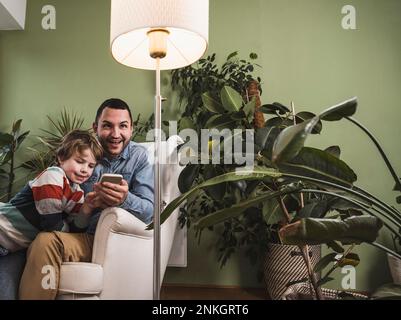 Happy son with father controlling light of floor lamp through smart phone Stock Photo