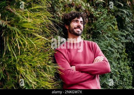 Thoughtful man with arms crossed leaning on plants Stock Photo
