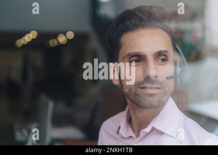 Smiling contemplative businessman wearing headset seen through glass Stock Photo