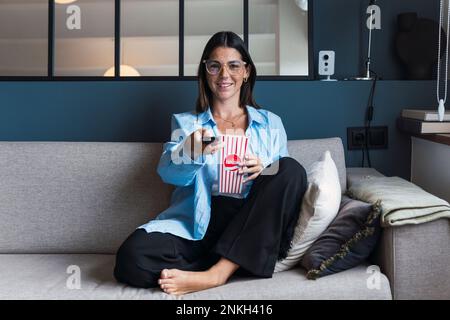 Smiling woman holding popcorn and TV remote control sitting on sofa at home Stock Photo