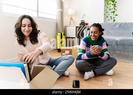 Smiling young woman unpacking box with friend holding coffee cup at home Stock Photo