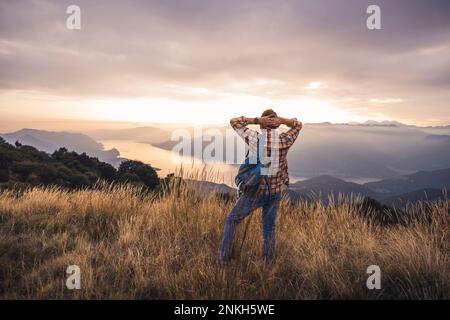 Mature man standing with backpack and hands behind head at sunset Stock Photo