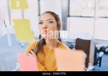 Thoughtful businesswoman with smart phone looking at adhesive notes on wall Stock Photo